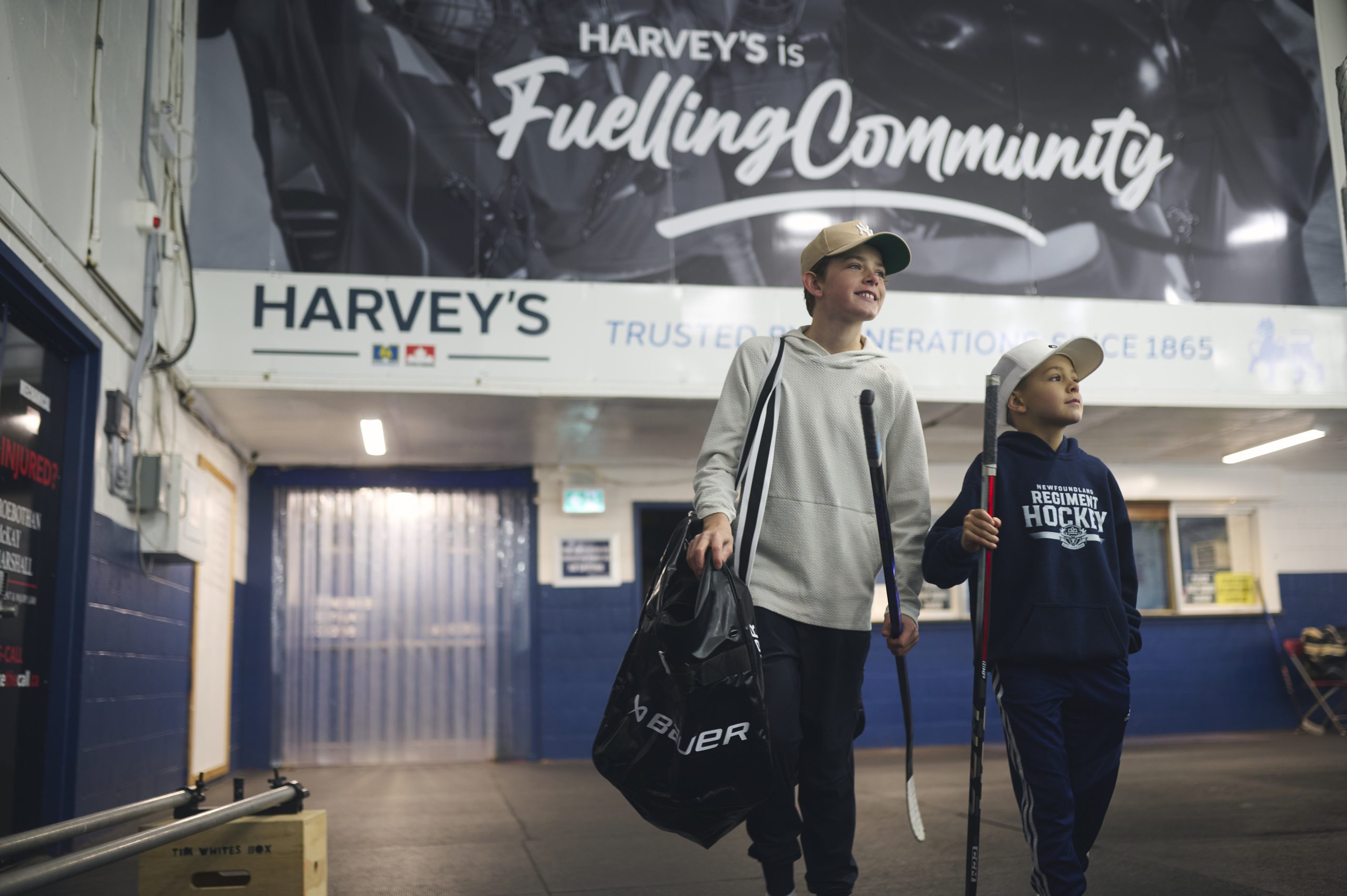 Two young boys walk through a hockey arena, carrying hockey sticks and an equipment bag, with the Harvey's Fuelling Community branding displayed on a wall behind them.