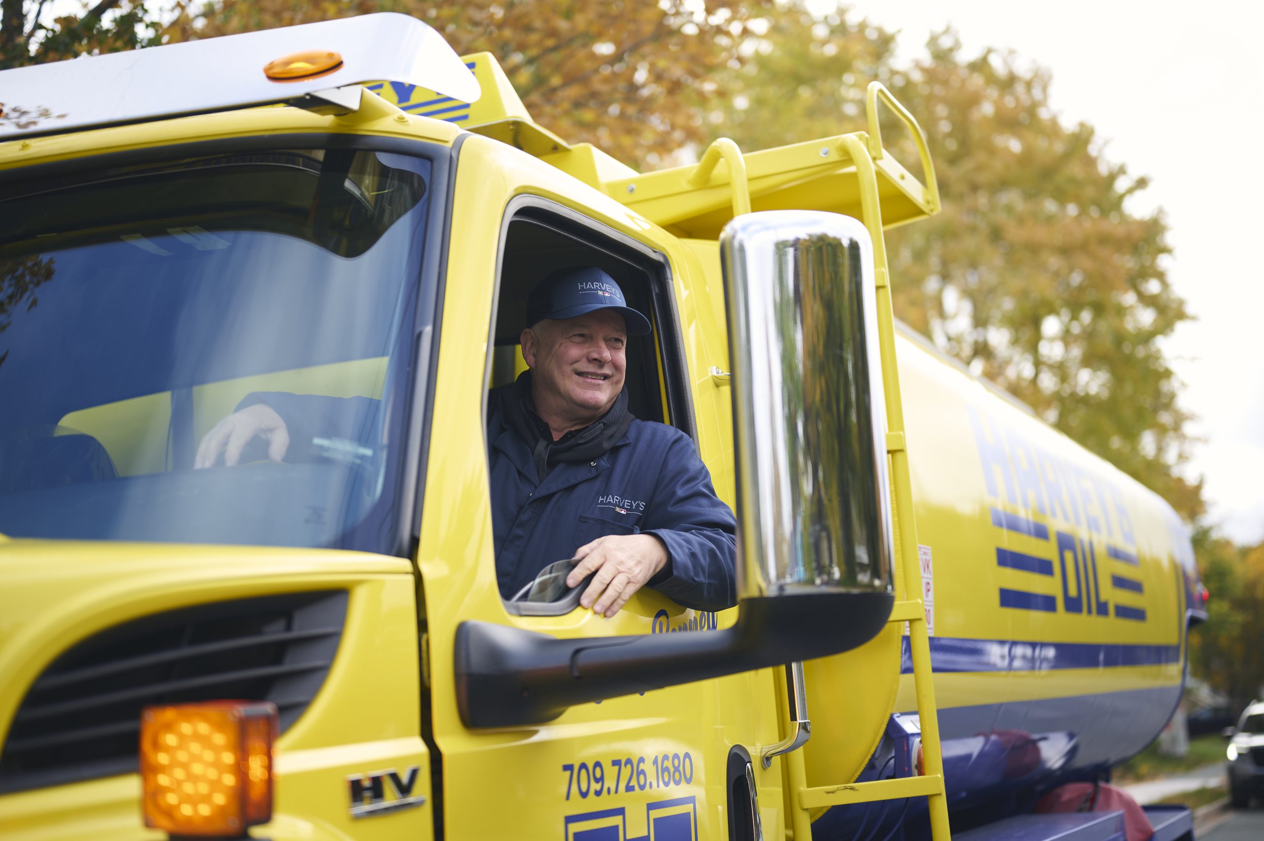 A Harvey's serviceman driving the retro-style fuel truck through a residential neighbourhood with a friendly smile.