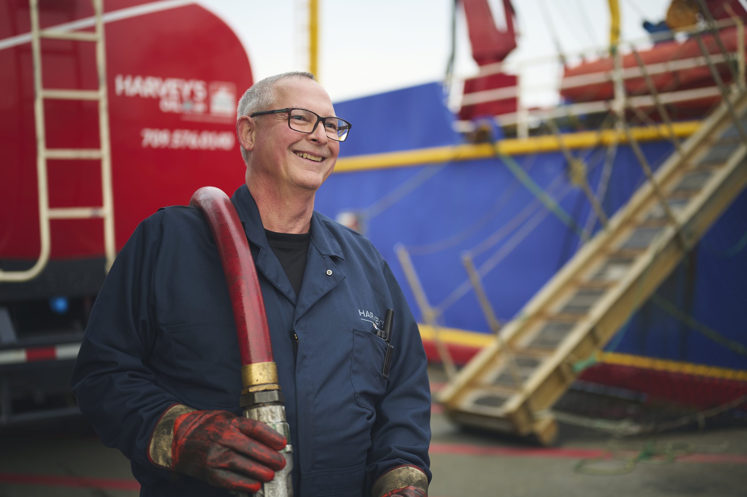 A smiling Harvey's journeyman in a shipyard, carrying the hose from a diesel delivery truck.