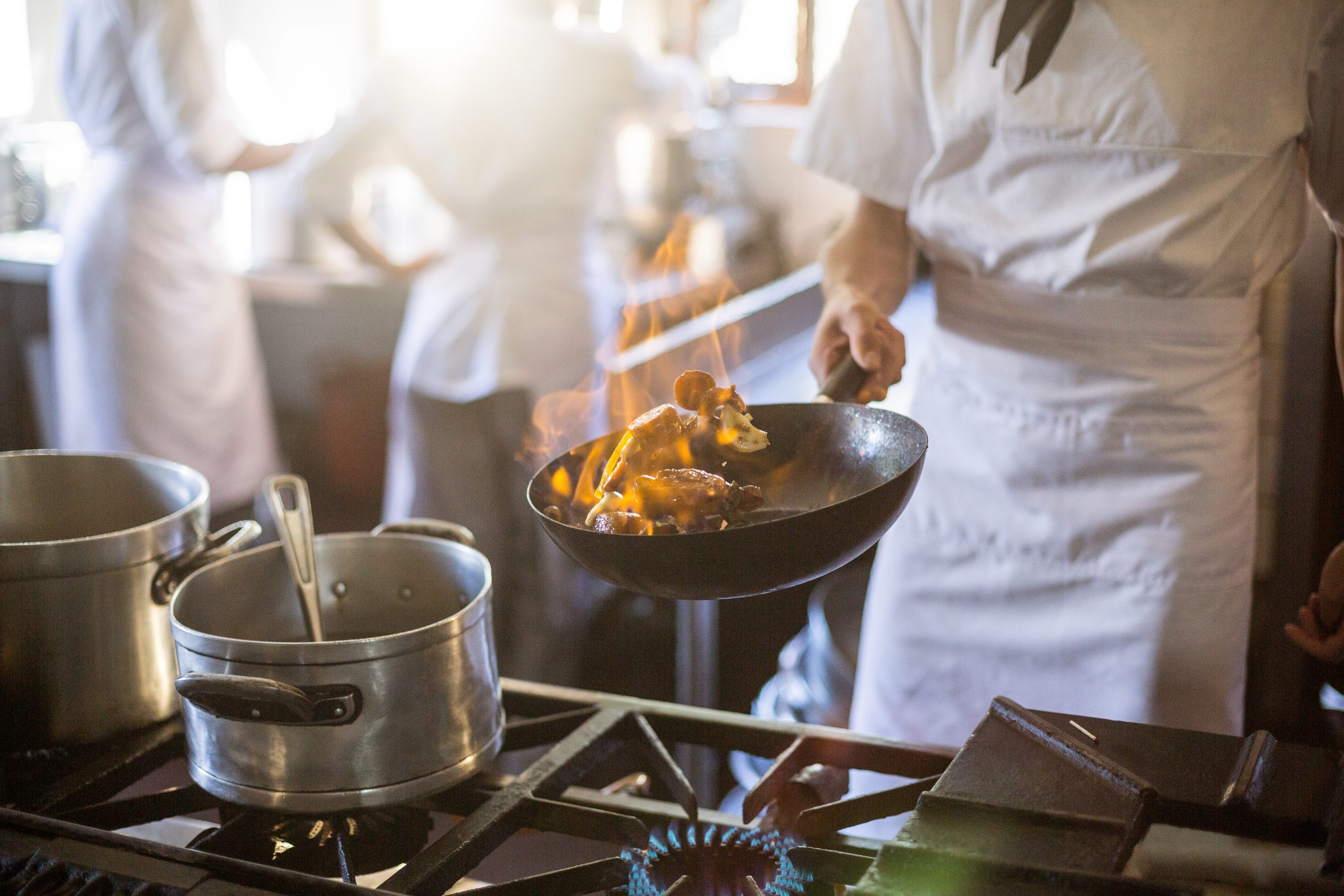 A chef in a restaurant kitchen, tossing stir fry in a pan over large flame.