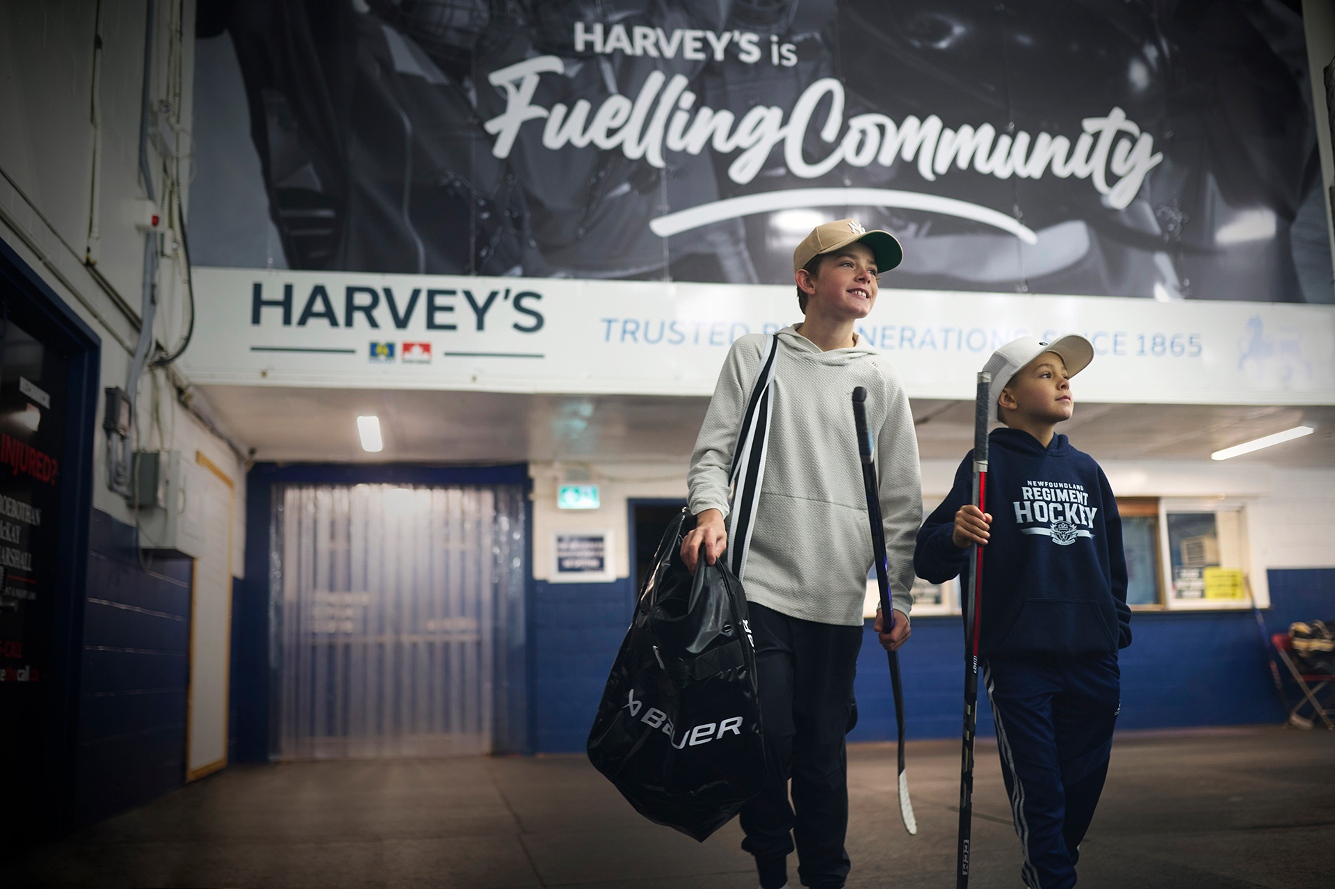 Two young boys walk through a hockey arena, carrying hockey sticks and an equipment bag, with the Harvey's Fuelling Community branding displayed on a wall behind them.