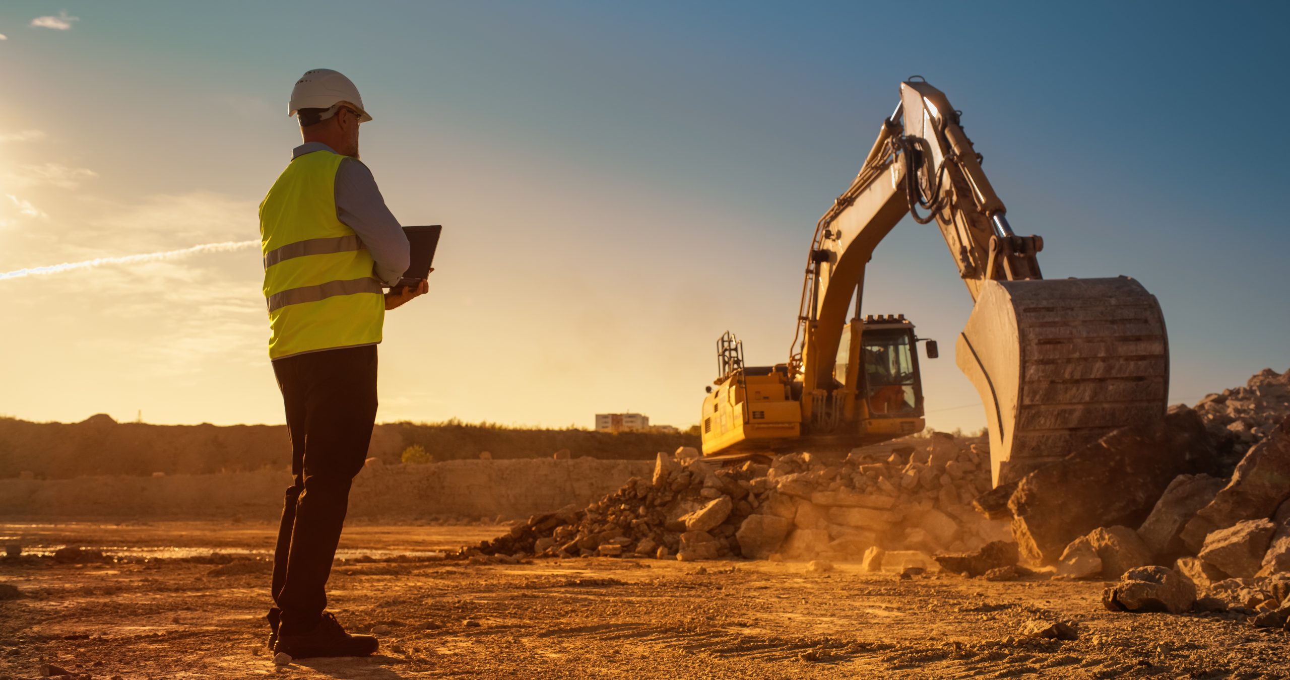 A civil engineer overseeing the operations of a backhoe at a construction site, and using a laptop.
