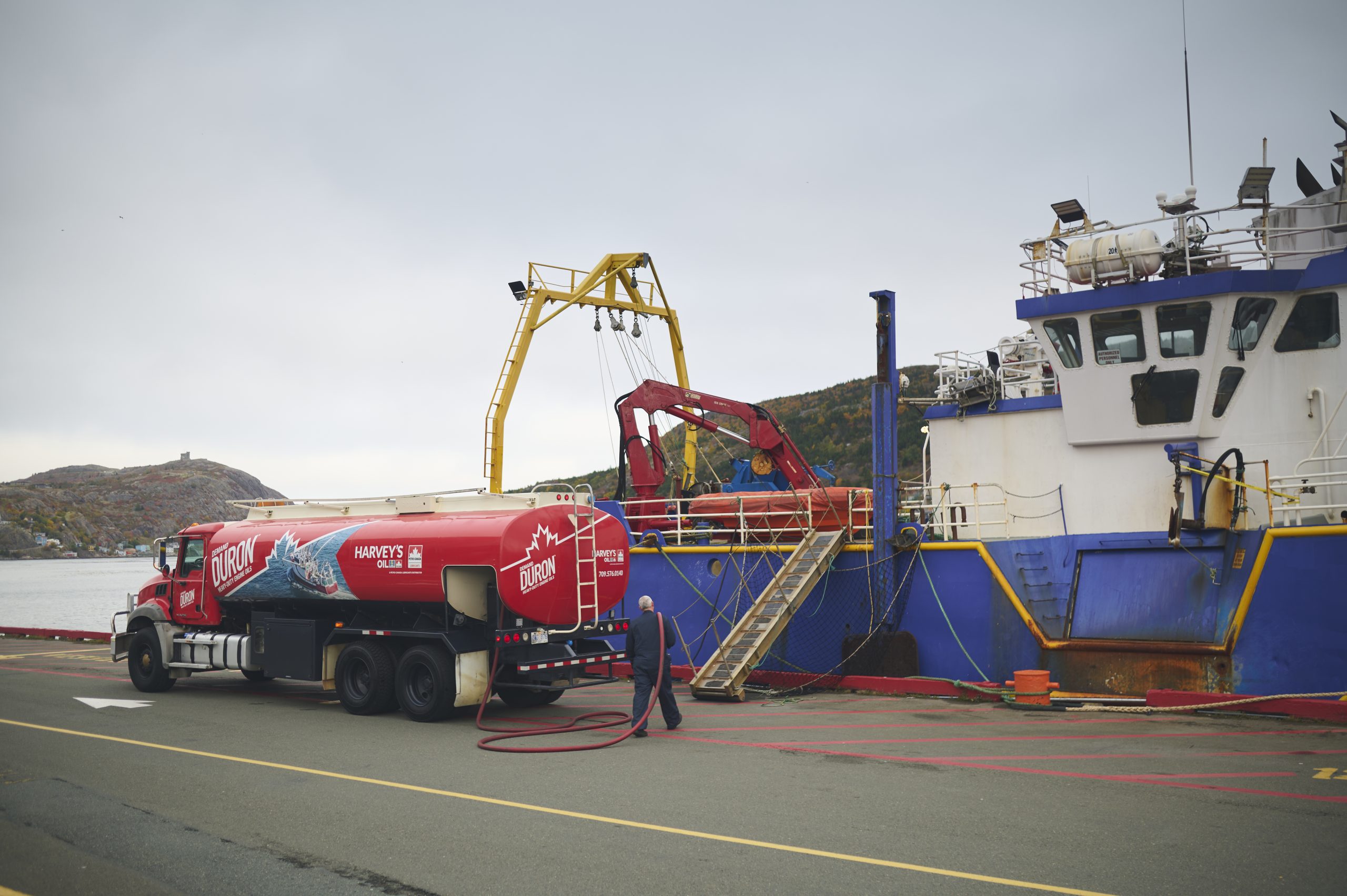 A Harvey's diesel truck parked on the St. John's harbour waterfront. A Harvey's journeyman carries the hose from the truck to a moored boat.
