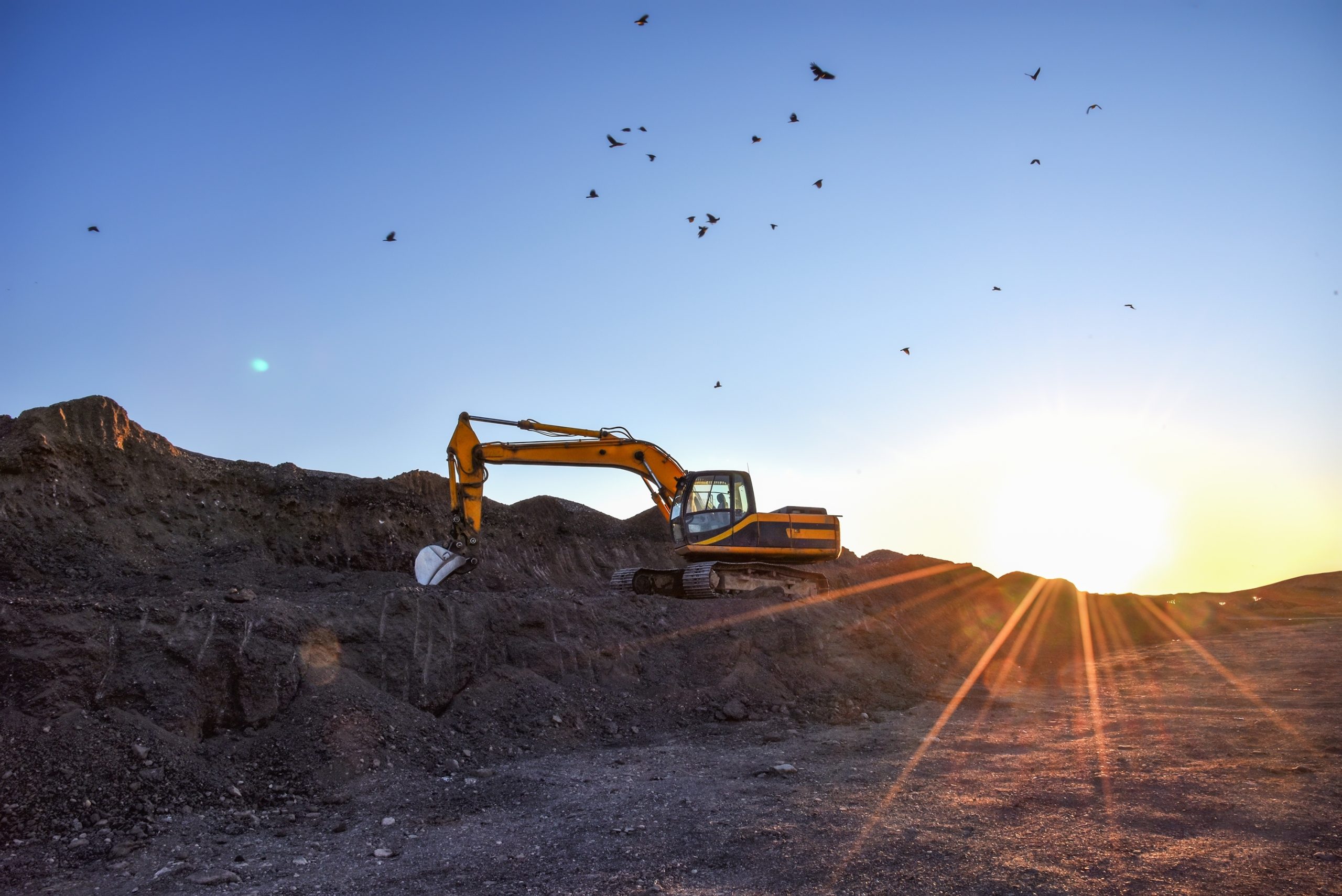 An excavator scooping up a large pile of dirt and soil at a construction site at sunset.