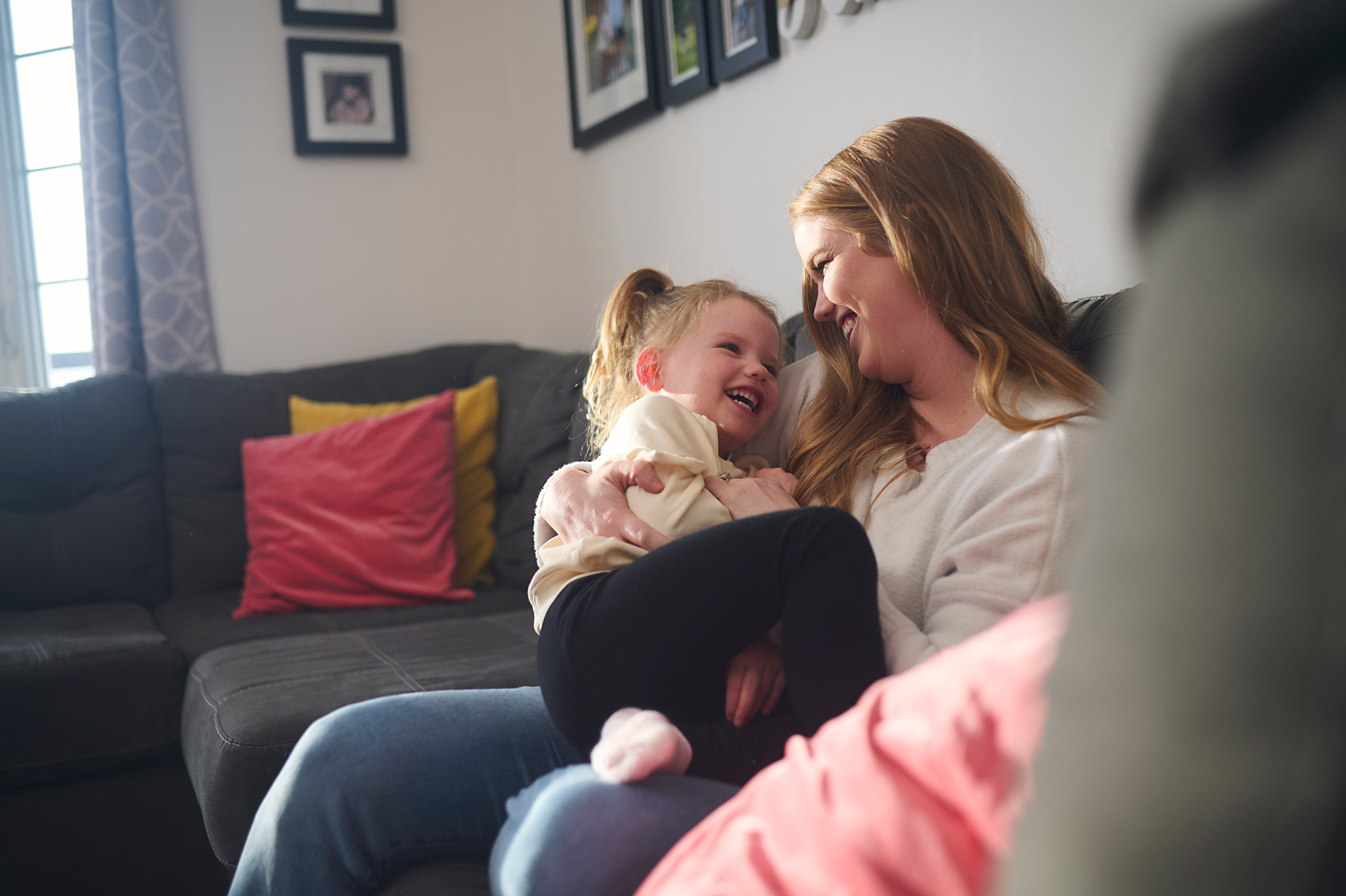 A mother and her young daughter snuggling and laughing on their living room couch.