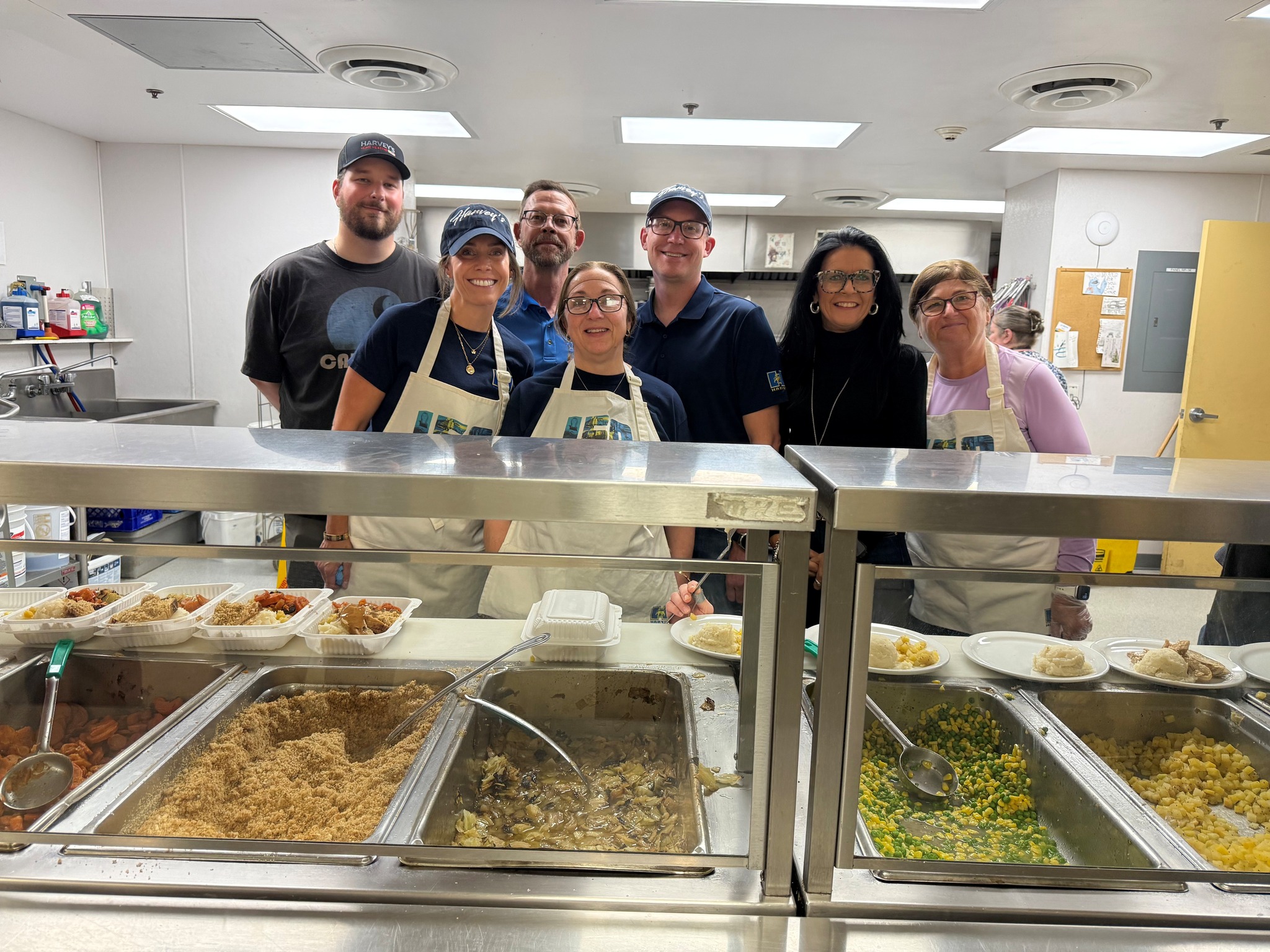 A group of smiling Harvey's employees in aprons, volunteering to feed the homeless in a soup kitchen or cafeteria-like setting.